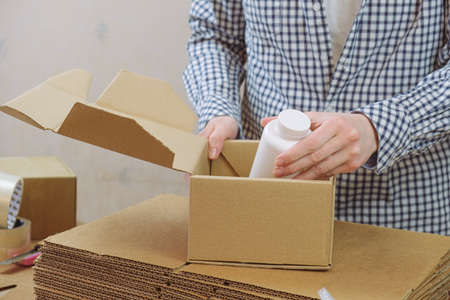 Woman in a warehouse is packing a plastic jar with vitamins in a cardboard box. Delivery of food supplements, vitamins and medicines. Online order processing, postage.の写真素材