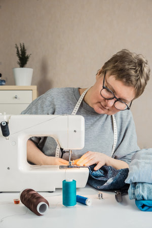 Woman with glasses sits at a table with a sewing machine and sewing supplies. A seamstress at her...