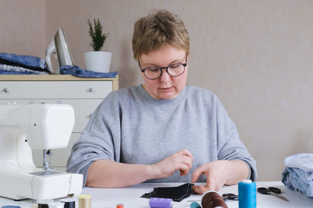 Woman with glasses sits at a table with a sewing machine and sewing supplies and makes a seam with a...