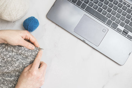 Close-up of female hands with knitting needles and knitting in front of a laptop on a light background. Online needlework classes, master class. The woman knits.の写真素材