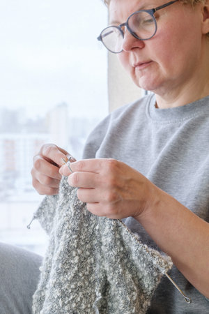 Middle-aged woman is knitting a gray wool scarf sitting by the window. The woman is doing...