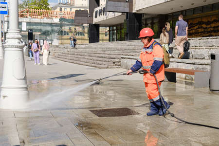 Woman from a city improvement service washes the sidewalk with water from a hose. City street cleaning service. Moscow, Russia - 05.21.2022のeditorial素材