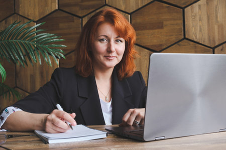 Attractive red-haired woman with shoulder-length hair sits at a desk with a laptop and a notepad. Portrait of a woman in a blue jacket in the office. Woman is looking at the camera.の写真素材