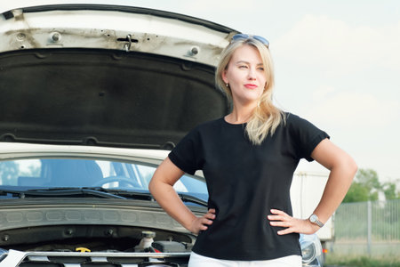 Confident blonde woman stands in front of a car with the hood up and looks away. Woman waiting for...