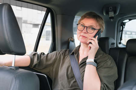 Business woman in glasses is talking on the phone while sitting in the back seat of a car. Business woman is negotiating while riding in a car. Serious middle aged woman with glassesの写真素材
