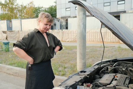 Confident blonde woman stands in front of a car with the hood up. Woman waiting for help on the road. Woman in a black blouse stands next to a broken down car.の写真素材