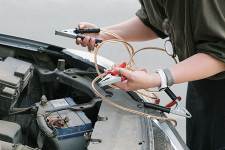 Woman stands in front of a car with the hood open and holds the battery charging clamps. Using jumper cables to charge dead car batteryの写真素材