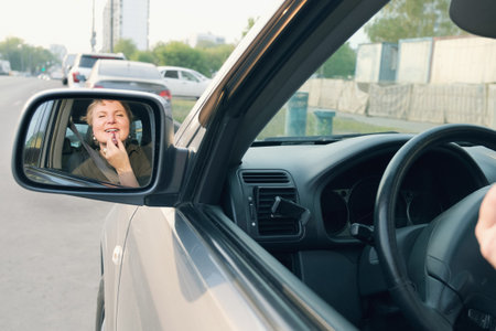 Middle aged woman with short hair driving a car paints her lips with lipstick. The woman driving...