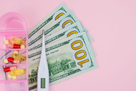 US dollar bills, an electronic medical thermometer and pills in a plastic box on a pink background....