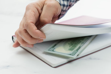 Close-up of a human hand opening a diary containing hundred dollar bills. The concept of giving a bribe, offering illegal rewards in order to achieve one's interests.の写真素材
