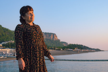 Mature woman in a dark summer dress stands on a pier near the sea against the backdrop of a landscape with mountains. A woman admires the sunset on the sea coast.の写真素材