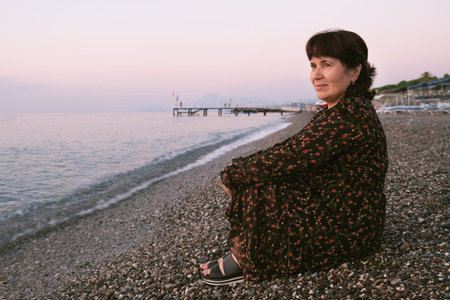 Elderly brunette woman sits at the edge of the sea and looks at the sunset. A woman on a pebble beach admires the sea. vacation in retirement.の写真素材