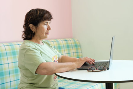 Mature brunette woman works in front of a laptop in a cafe. Elderly woman in profile in front of a...