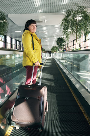 Vertical full length portrait of an elderly woman with a suitcaseat the airport. Mature brunette woman at the airport stands on a moving walkway. Woman in the departure area of international airportの写真素材