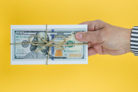 Man holding in the palm of his hand white paper envelope with a hundred dollar bill tied with twine on a bright yellow background. The concept of money as a gift, bribe, postal itemの写真素材
