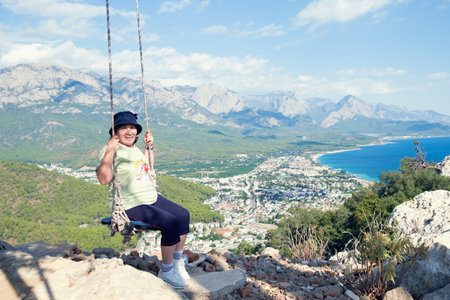 Mature woman in sportswear sitting on makeshift swing on a rope twine over an observation deck in the mountains. Beautiful view from the mountain to the city at the foot. Tourist extreme attraction.の写真素材