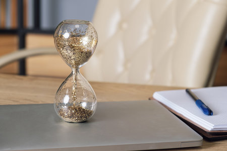 Glass hourglass stands on a laptop on a desk. Desktop with a laptop, notepad and pen on the...