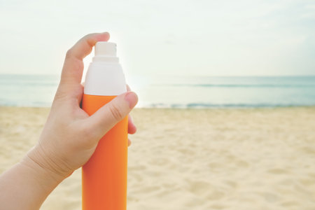Womans hand holds a sunscreen spray in an orange bottle against the backdrop of a sandy beach and...