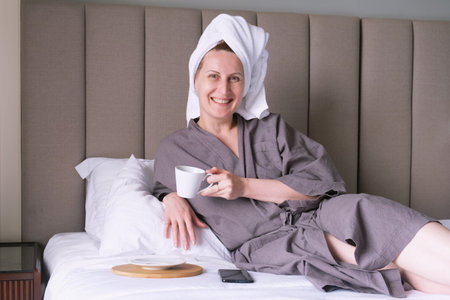 Middle-aged woman with a towel on her head drinks coffee in bed. A woman is resting in a hotel and...
