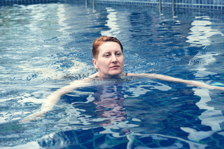Middle-aged woman swims in the pool and smiles. Beautiful woman with short red hair is relaxing in...