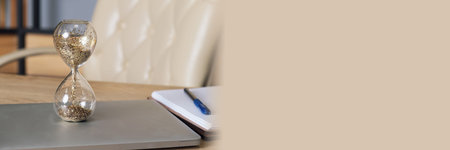 Glass hourglass stands on a laptop on a desk. Desktop with a laptop, notepad and pen on the...
