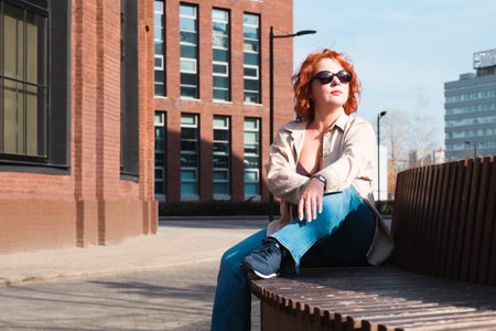 Attractive red-haired woman with sunglasses sits on a bench in a business district. Woman on a spring walk. Confident middle-aged woman.の写真素材