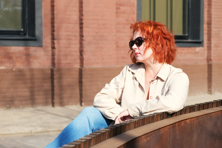 Attractive red-haired woman with sunglasses sits on a bench in a business district. Woman on a spring walk. Confident middle-aged woman.の写真素材