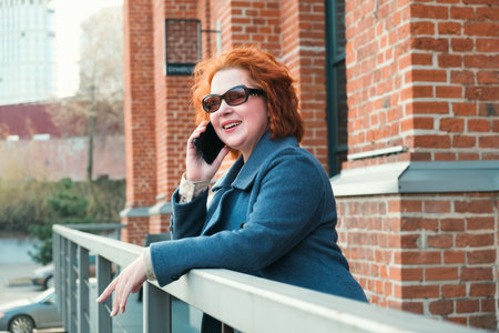 Attractive red-haired woman in a blue coat is talking on a mobile phone. Woman smiling and gesturing emotionally during a telephone conversation.の写真素材