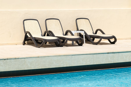 Three sun loungers and a plastic table at the edge of the hotel pool. Outdoor swimming pool with handrails and empty sunbeds at resort on sunny dayの写真素材
