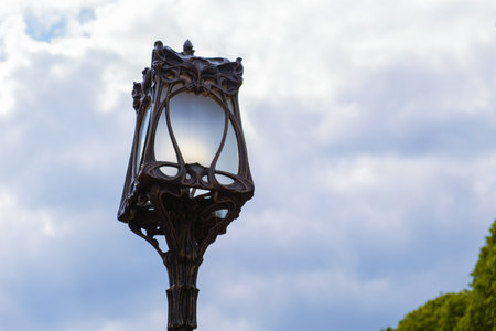 Iron openwork street lamp with a transparent glass shade against a cloudy sky. Street lighting, urban illumination, improvement of the city.の写真素材