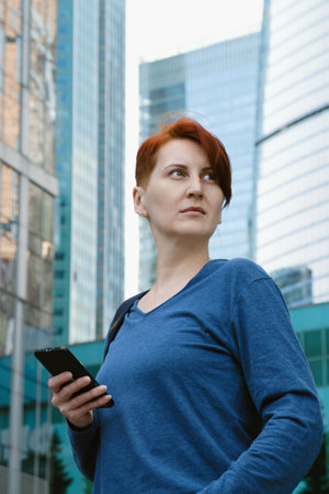 Middle-aged woman in a blue shirt stands in a modern business district with a smartphone in her hand...