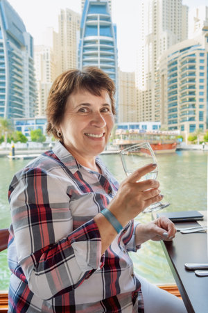 Senior citizen woman sits at a table on the deck of a pleasure boat and drinks water and looks at the camera. Woman on a boat ride in Dubai Marina. Retired woman tourist on an excursion.の写真素材