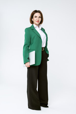 Attractive middle-aged business woman in black trousers and a green jacket stands with a laptop on a white background. Full-length portrait of a business lady.の写真素材