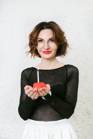 Attractive middle-aged woman holds a red cake with a birthday candle in her palms. Symbol of the celebration of a birthday, anniversary, anniversary.の写真素材