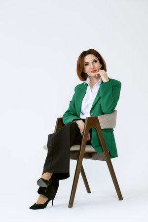 Attractive middle-aged business woman in black trousers and a green jacket is sitting on a chair on a white background. Portrait of a business lady.の写真素材