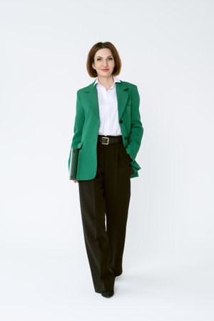 Attractive middle-aged business woman in black trousers and a green jacket stands with a laptop on a white background. Full-length portrait of a business lady.の写真素材