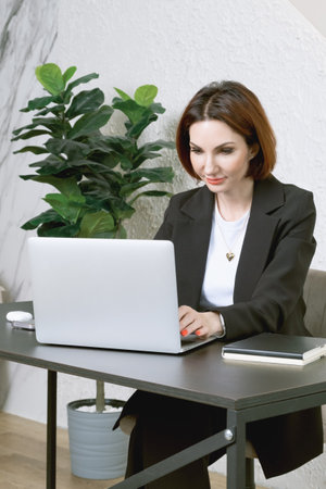 Portrait of business woman in a black business suit at desk in your office. Business portrait. Business woman is sitting in front of laptop screenの写真素材