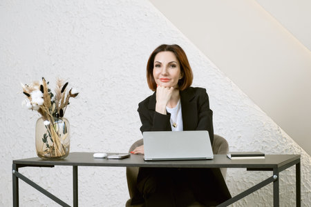 Portrait of business woman in a black business suit at desk in your office. Business portrait. Business woman is sitting in front of laptop screenの写真素材