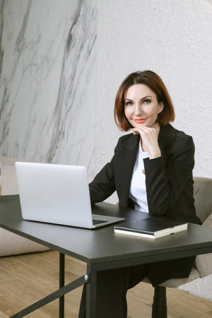 Portrait of business woman in a black business suit at desk in your office. Business portrait. Business woman is sitting in front of laptop screenの写真素材