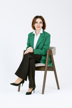 Attractive middle-aged business woman in black trousers and a green jacket is sitting on a chair on a white background. Portrait of a business lady.の写真素材