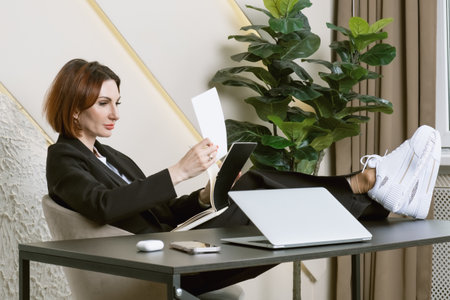 Portrait of a business woman in a black suit at her desk in the office. A woman in a relaxed position is studying documents with her feet on the table.の写真素材