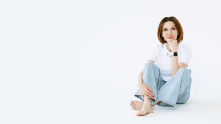 Attractive middle-aged woman in jeans and barefoot on a white background. Portrait of a woman in casual clothes. A banner with an empty space for textの写真素材