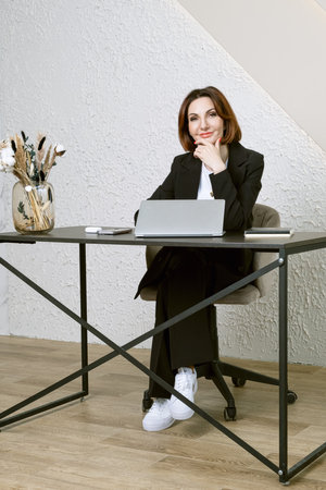 Portrait of business woman in a black business suit at desk in your office. Business portrait. Business woman is sitting in front of laptop screenの写真素材