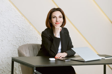 Portrait of business woman in a black business suit at desk in her office. Business portrait. Business woman is sitting in front of laptop screenの写真素材