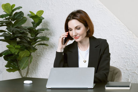 Portrait of businesswoman in the black jacket at desk in your office. Business portrait. Business woman is sitting in front of laptop screen and talking on cell phoneの写真素材