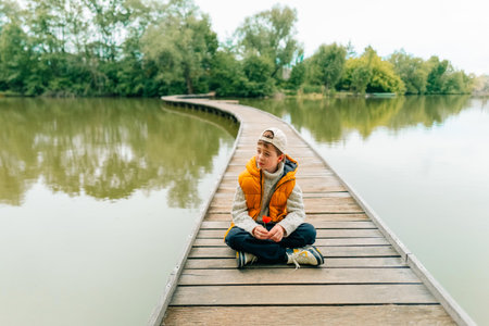 A teenage boy sits on a bridge on a pond and holds a poppy flower in his handsの写真素材