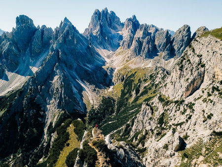 Rugged dolomite mountains rise against a clear blue sky, highlighting diverse rock formations and lush greenery in the valleys below. A hiker explores this stunning landscape.の写真素材