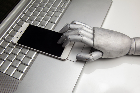 Glossy smartphone with robot hand touching it by finger on laptopの写真素材