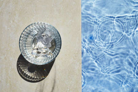 A glass of iced cold water placing on the travertine marble stone by the beautiful swimming pool in the sunny morning.の写真素材