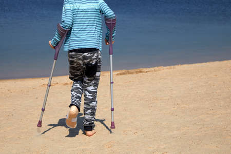 A little boy with Autism, Asperger syndrome on crutches after breaking his leg walks on the beach.の写真素材